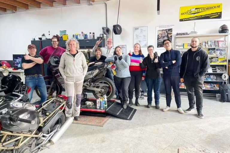 Groep mensen poseert in een garage met motoren en gereedschappen.