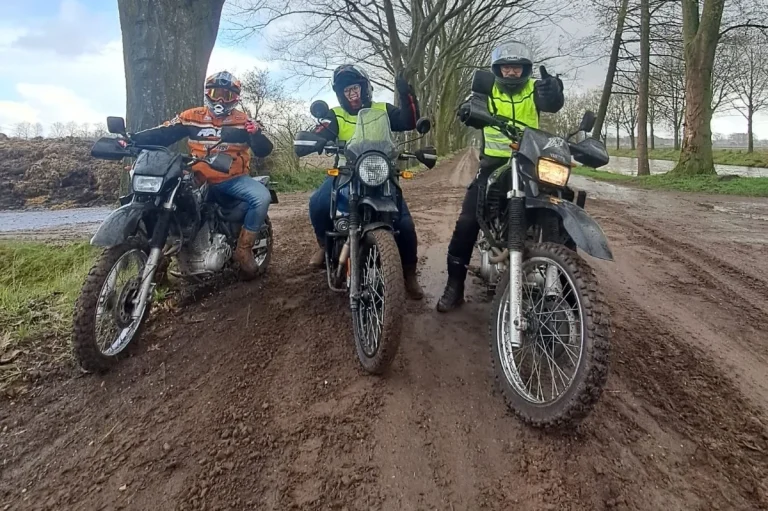Drie motorrijders op offroad motorfiets staan op een modderige weg tussen bomen.