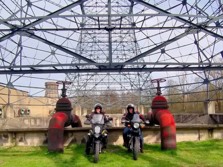 Twee motorrijders staan voor grote, rode waterkranen onder een metalen structuur.